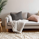 **

"A cozy living room bathed in soft, natural light. White walls, a light gray sofa with chunky knit throw blankets, and several cushions in muted pastel colors. A sheepskin rug lies on the floor near a birch wood coffee table. Candles are lit on the table, and a snake plant sits in a stylish pot on a nearby shelf. The overall feeling is warm, inviting, and 'hygge'."

**